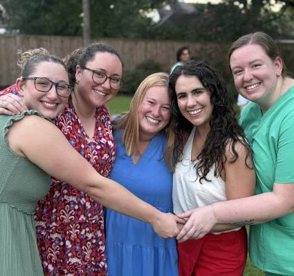 Five people standing outdoors on a grassy lawn with arms around one another and hands joined in the center. The group is wearing colorful clothing, including green, patterned red, blue, white, and mint-green outfits. A wooden fence, trees, and shrubs are visible in the softly blurred background, with another person out of focus in the distance. Natural outdoor lighting and a shallow depth of field keep the group as the focal point of the image.