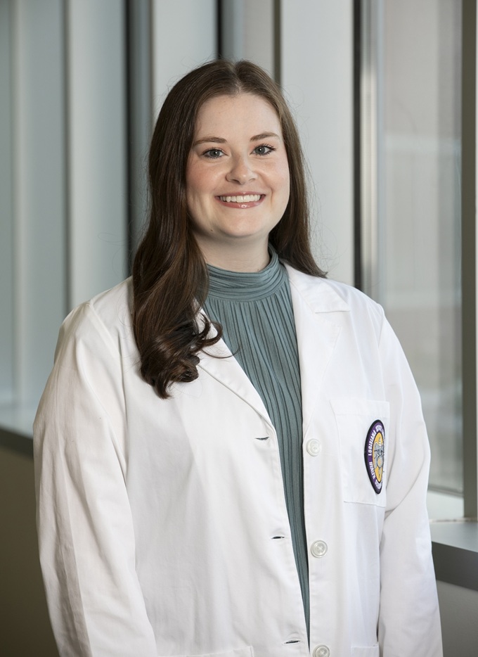 Professional clinical portrait of a person standing indoors near large windows, wearing a white medical coat over a high-neck, pleated sage-green top. The subject faces the camera in a formal pose. A Louisiana State University Health Sciences Center patch is visible on the coat pocket, and the background consists of softly blurred vertical architectural elements and natural light.