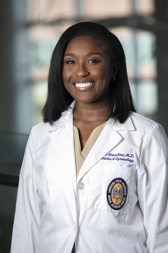 Professional portrait of a person wearing a white medical coat over a beige top, photographed indoors against a softly blurred background with large windows. The coat features purple embroidered text indicating a specialty in Obstetrics & Gynecology and a Louisiana State University Health Sciences Center patch on the pocket. The subject is facing the camera in a formal head-and-shoulders pose.