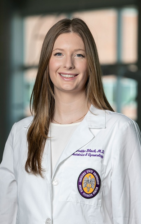 Professional head-and-shoulders portrait of a person wearing a white medical coat and light-colored top, standing indoors against a softly blurred background with large windows. The coat features embroidered text indicating a medical specialty and a Louisiana State University Health Sciences Center emblem on the front pocket. Long straight hair falls over the shoulders, and the subject faces the camera.