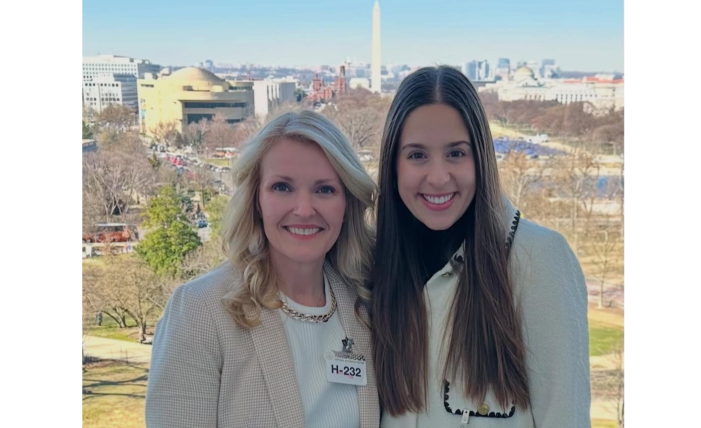 Two people stand side by side on an outdoor terrace overlooking the National Mall in Washington, D.C. Both are wearing visitor badges labeled “H-232.” The person on the left wears a light-colored blazer over a white top, and the person on the right wears a light-colored jacket over a dark outfit. In the background, the Washington Monument rises above trees and government buildings under a clear blue sky. The photo is taken during daylight from an elevated vantage point, with the city skyline visible in the distance.