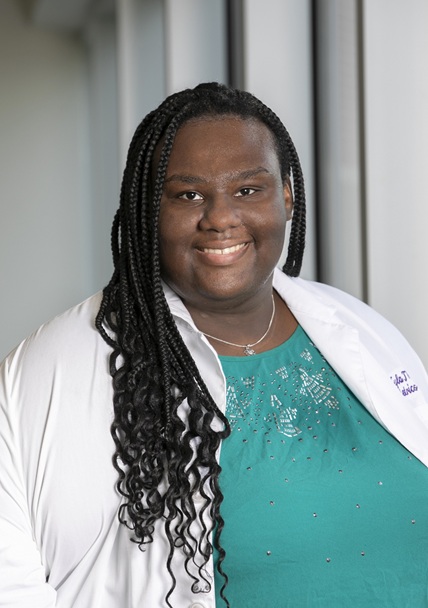 Professional clinical portrait of a person standing indoors near large windows, wearing a white medical coat over a teal top with decorative bead and rhinestone accents. The subject faces the camera in a formal pose. The white coat includes partially visible embroidered text and appears to be associated with a medical training program. The background features softly blurred vertical window frames and natural light.
