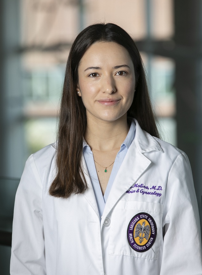 Professional head-and-shoulders portrait of a person wearing a white medical coat over a light blue collared shirt, photographed indoors against a softly blurred background with large windows. The coat features purple embroidered text indicating a specialty in Obstetrics & Gynecology and a Louisiana State University Health Sciences Center patch on the front pocket. The subject faces the camera in a formal clinical portrait setting and wears a delicate necklace with a small green pendant.