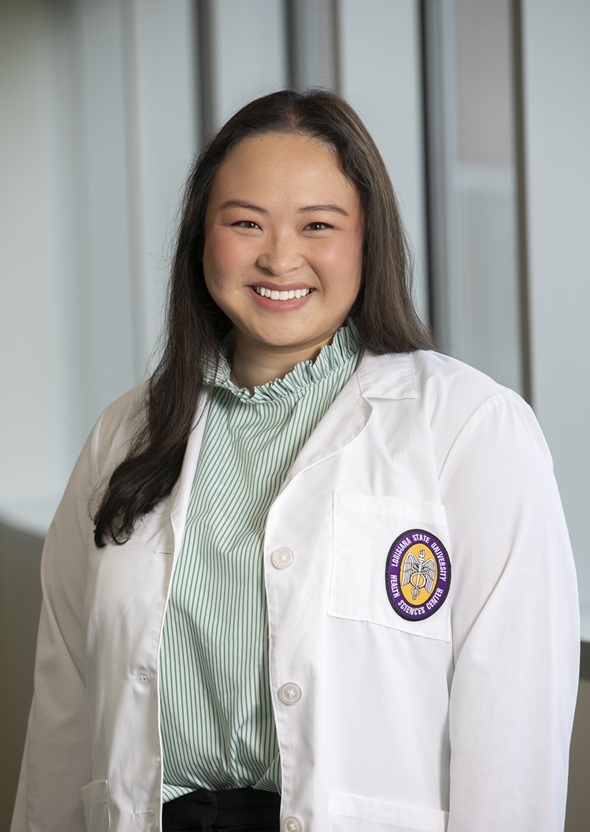 Professional clinical portrait of a person standing indoors near large windows, wearing a white medical coat over a green-and-white striped blouse with a ruffled mock neck. The subject faces the camera in a formal pose. A Louisiana State University Health Sciences Center patch is visible on the coat pocket. The background features softly blurred vertical architectural elements and natural light, creating a clean healthcare setting.