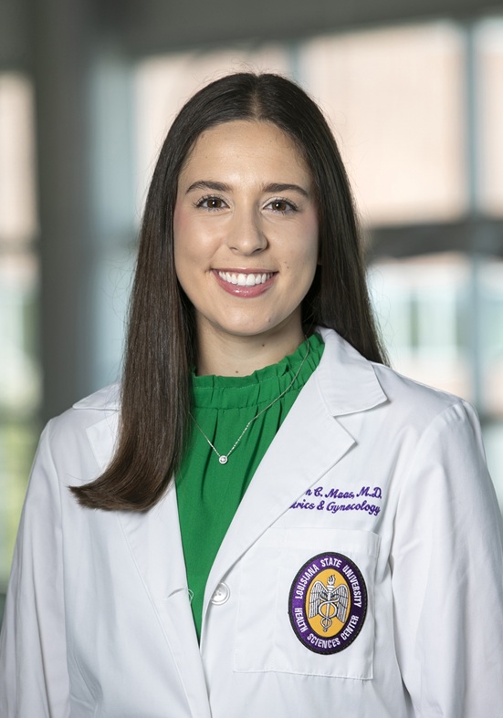 Professional head-and-shoulders portrait of a person wearing a white medical coat over a bright green blouse, photographed indoors against a softly blurred background with large windows. The white coat features purple embroidered text indicating a specialty in Obstetrics & Gynecology and a Louisiana State University Health Sciences Center patch on the front pocket. The subject faces the camera in a formal clinical portrait setting and wears a small pendant necklace.