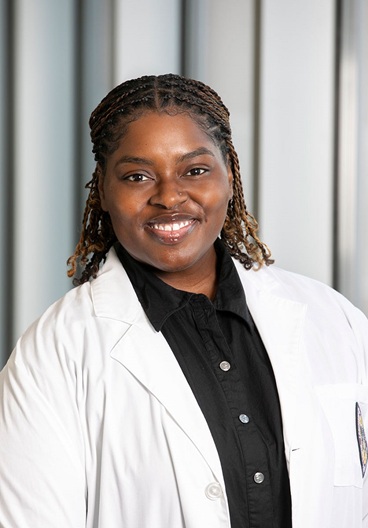 Professional head-and-shoulders portrait of a person wearing a white medical coat over a black button-up shirt, photographed indoors against a softly blurred background with vertical architectural panels. The subject faces the camera in a formal clinical portrait setting. The white coat includes a partially visible institutional patch on the pocket, and the person has braided hair styled away from the face.