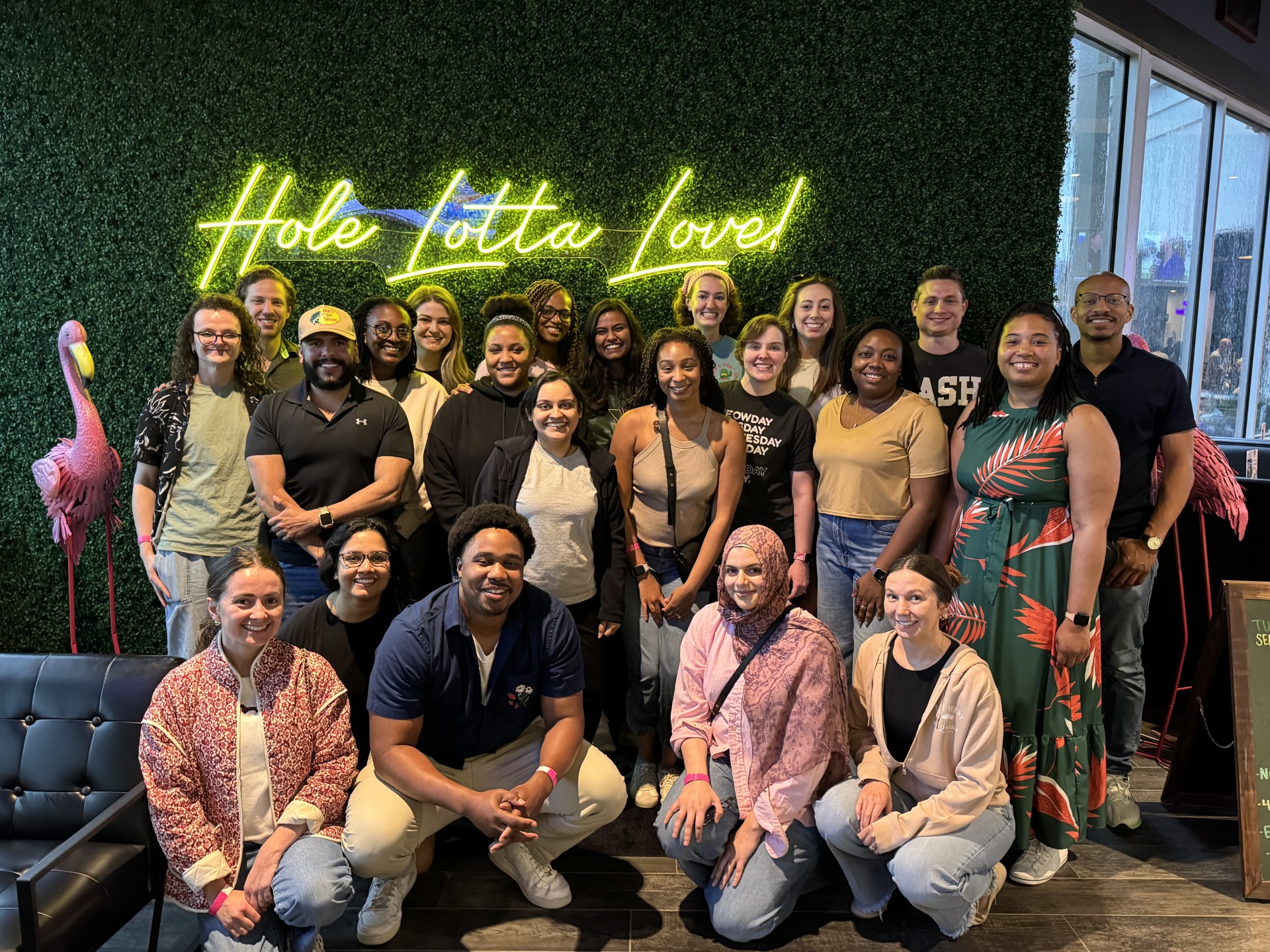 A large group of people pose together indoors in front of a glass wall and industrial‑style interior. Several people in the back row hold bright green rectangular signs with large purple letters that spell “LSUHSC.” The group is arranged in rows, with some people standing and others crouching in front. The space has dark flooring, exposed structural elements, and natural light coming through the glass behind the group.