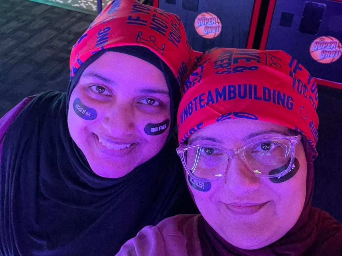 Close-up selfie of two individuals at an indoor entertainment venue with colorful purple and blue arcade lighting. Both participants are wearing red team bandanas with printed event graphics and black sticker strips on their cheeks displaying phrases such as “Bring It On,” “High Fiver,” “Winner,” and “Boom.” Arcade basketball game machines are visible in the background. The image captures a team-building or wellness event in a lively arcade setting.