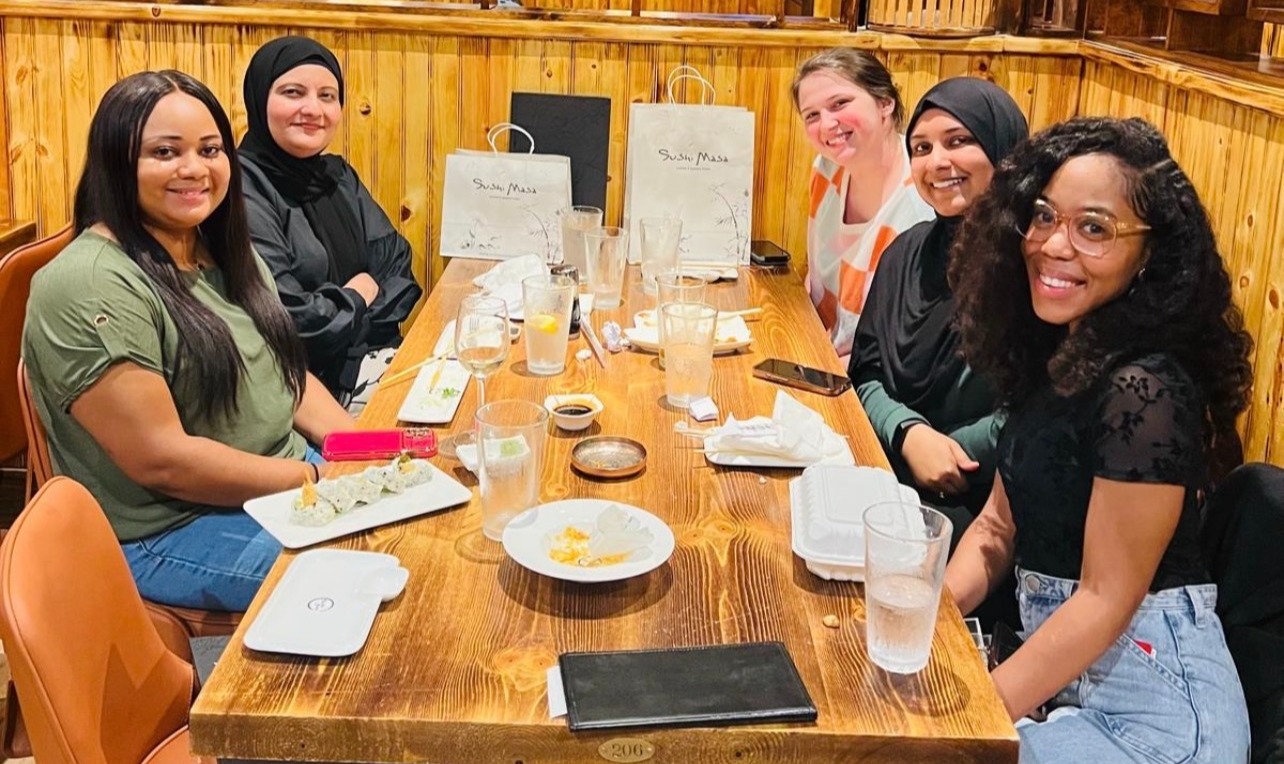Six people are seated around a large wooden table at a restaurant, posing for a group photo during a shared meal. Plates, glasses, takeout containers, and sushi dishes are arranged on the table, with several shopping or takeaway bags visible in the background. The restaurant features warm wood-paneled walls and booths, creating a casual dining atmosphere. The group is gathered together for a social wellness or team-building event.