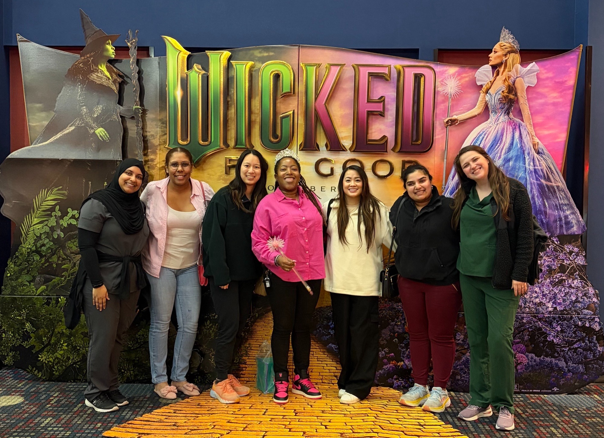 Seven individuals pose together in front of a large Wicked movie display inside a theater or entertainment venue. The backdrop features the title “WICKED” and life-size promotional images of two characters standing on either side of a yellow brick road graphic. One person in the center wears a tiara and holds a wand, while the others are dressed in casual clothing and scrubs. The group stands on the yellow brick road design in front of the colorful movie-themed backdrop, creating a commemorative photo from a wellness or team outing event.