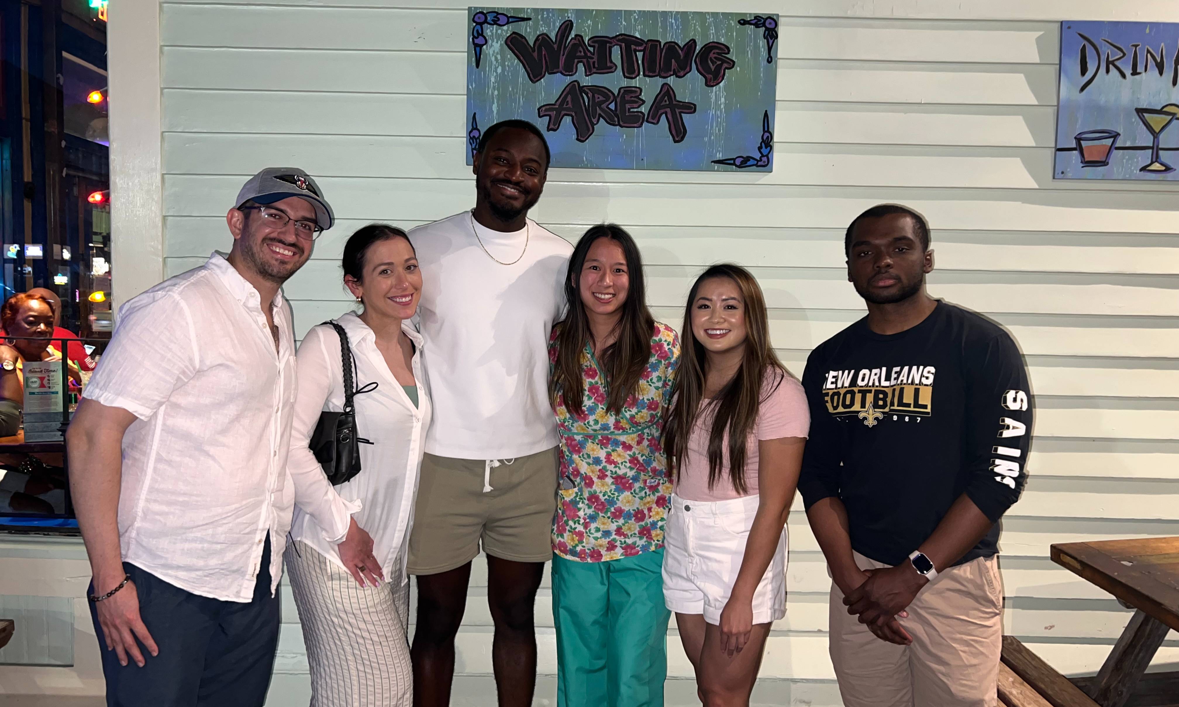 Six people pose together indoors in front of a wall with a sign reading “Waiting Area.” The group is dressed in a mix of casual clothing and scrubs, standing side by side for a group photo. The setting appears to be a restaurant or entertainment venue, with wooden picnic-style tables, string lights, and visible seating areas in the background. Additional signage, including a partially visible “Drink” sign, hangs on the wall nearby. The photo captures a social gathering or wellness event in a casual evening setting.