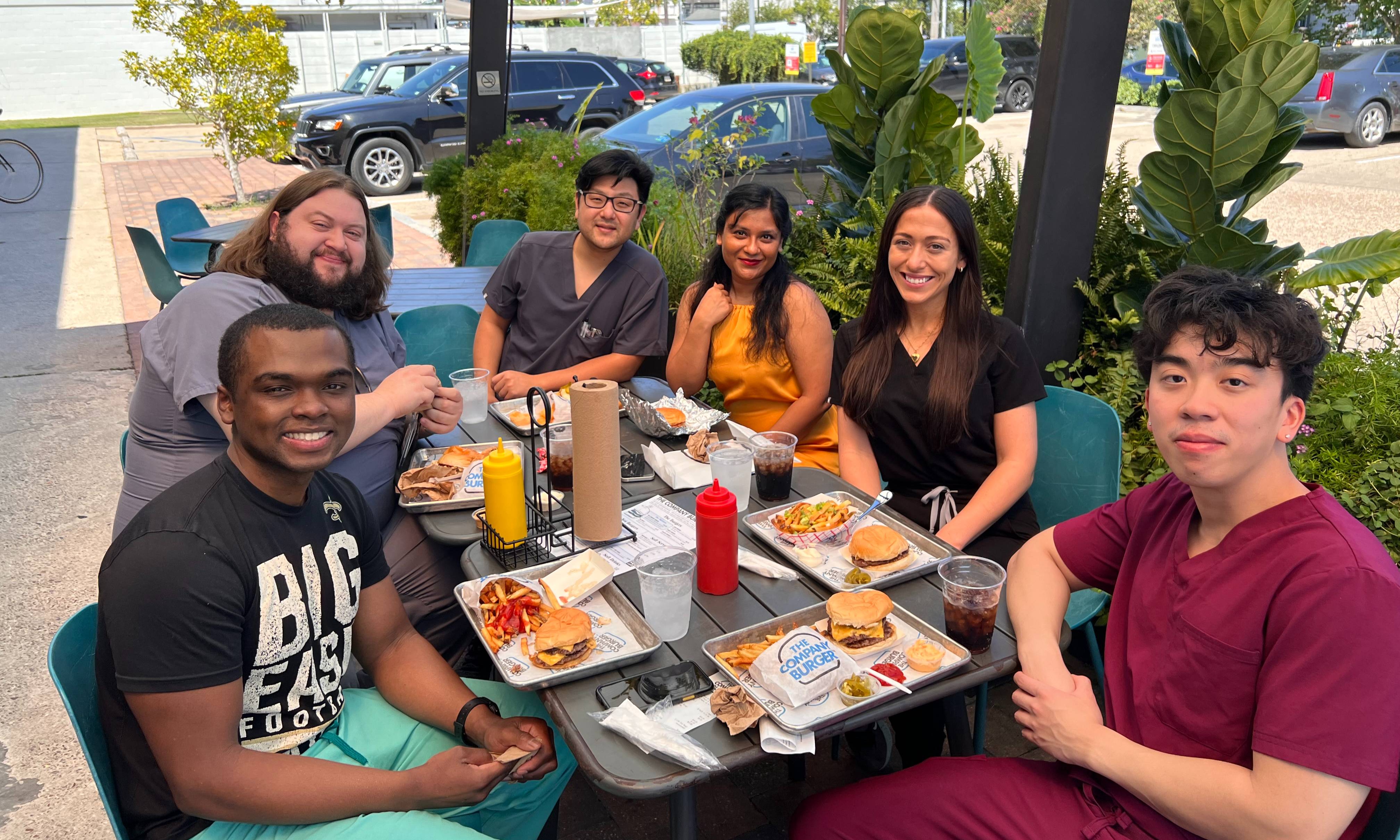 Six individuals are seated around an outdoor restaurant table during a group meal. Several people are wearing scrubs, and plates of burgers, fries, sandwiches, and drinks are arranged on the table alongside condiment bottles. The group is gathered on a covered patio surrounded by large green plants and hanging ferns. Parked cars, nearby buildings, and a sunny blue sky are visible in the background, creating a casual and social setting for a wellness or team-building event.