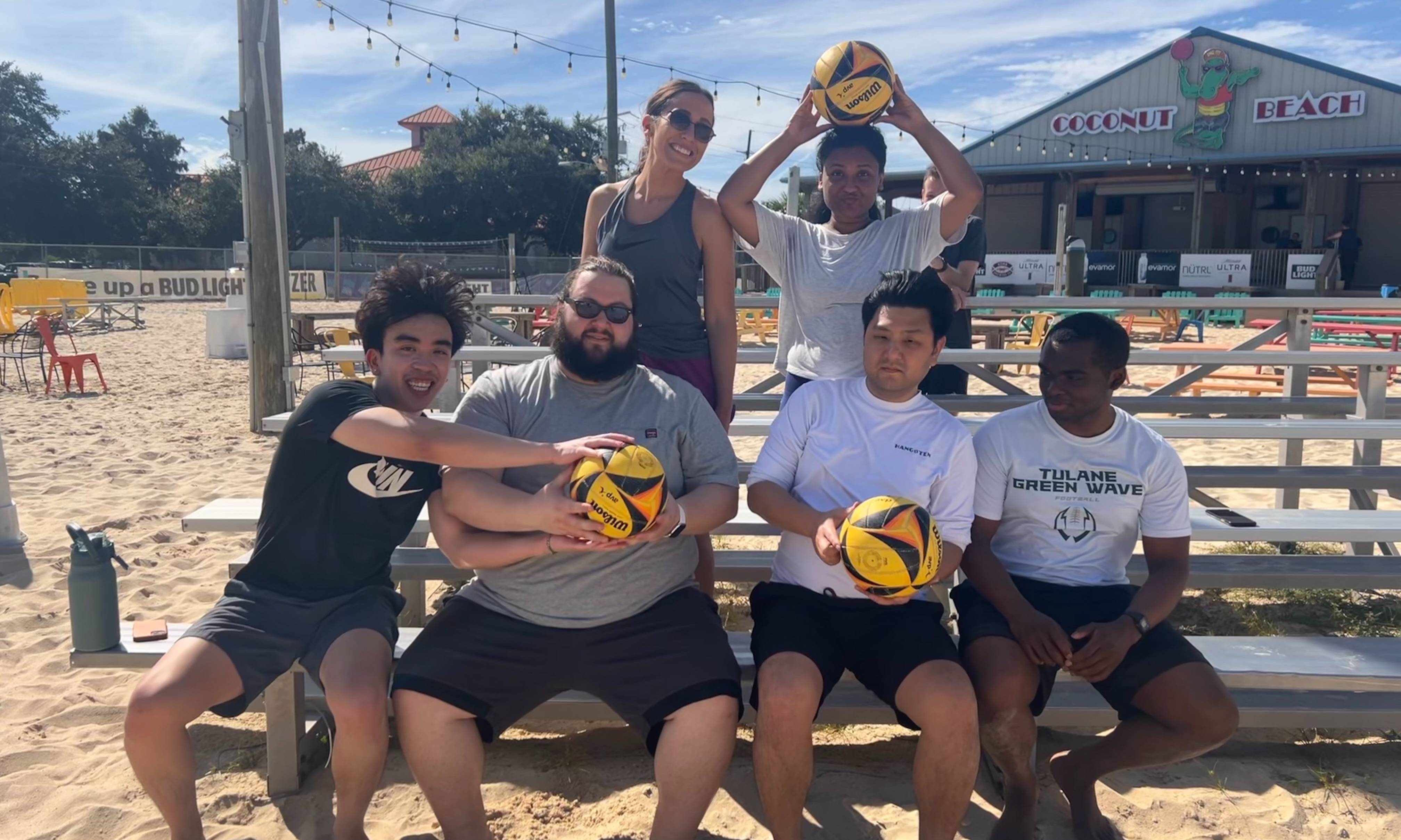 Six people pose together on outdoor bleachers at a sand volleyball venue, several holding yellow-and-black volleyballs. The group is dressed in casual athletic clothing and sits or stands in front of a sandy court with string lights overhead. A building with a sign reading “Coconut Beach” is visible in the background, along with picnic tables and volleyball court facilities. The photo captures a recreational wellness or team-building outing on a bright, sunny day with blue skies and scattered clouds.