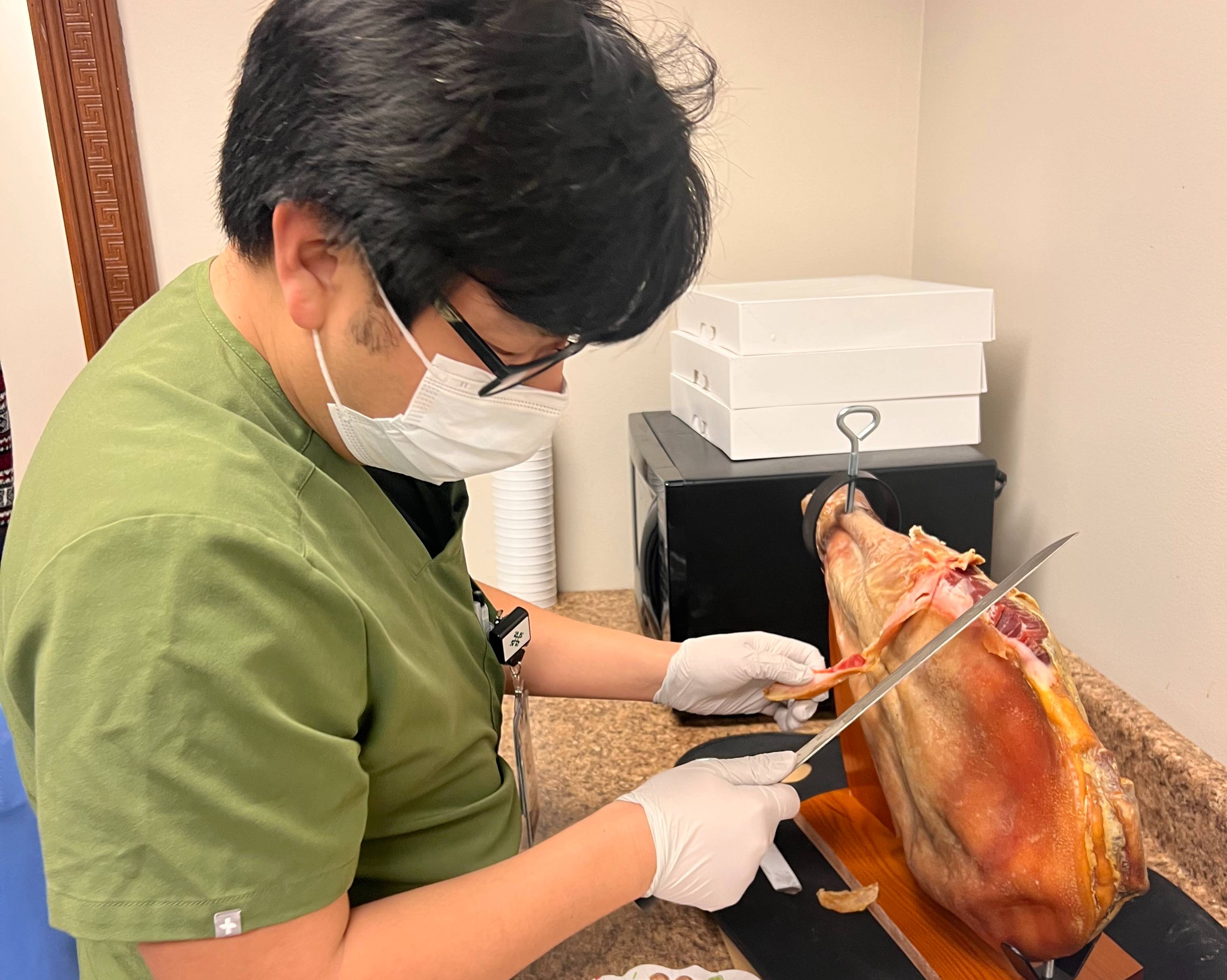 A person wearing green scrubs, gloves, eyeglasses, and a face mask slices a cured ham mounted on a wooden carving stand in a break room or kitchen area. The ham is secured on a countertop beside a microwave and stacked white boxes. Using a long carving knife, the person carefully cuts thin slices from the ham. The indoor setting and food preparation suggest a workplace celebration, holiday gathering, or wellness event.