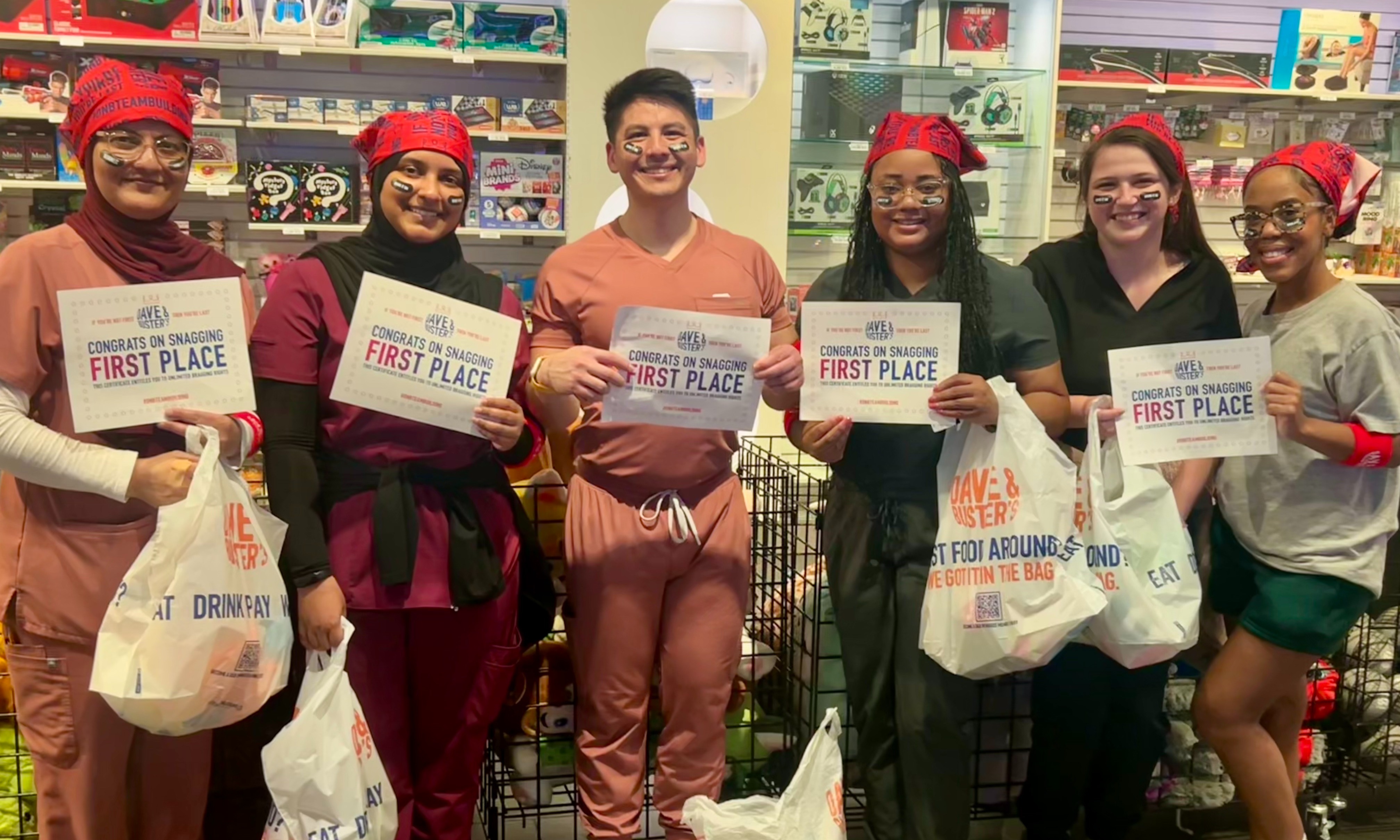 Six participants stand together inside an entertainment venue, holding certificates that read “Congrats on Snagging First Place” and carrying prize bags from Dave & Buster’s. Several participants are dressed in scrubs and wear red bandanas and black face-paint stripes as part of a team competition or wellness event. Shelves stocked with games, electronics, and prizes are visible in the background. The group is posed for a celebratory photo, highlighting a first-place team achievement.