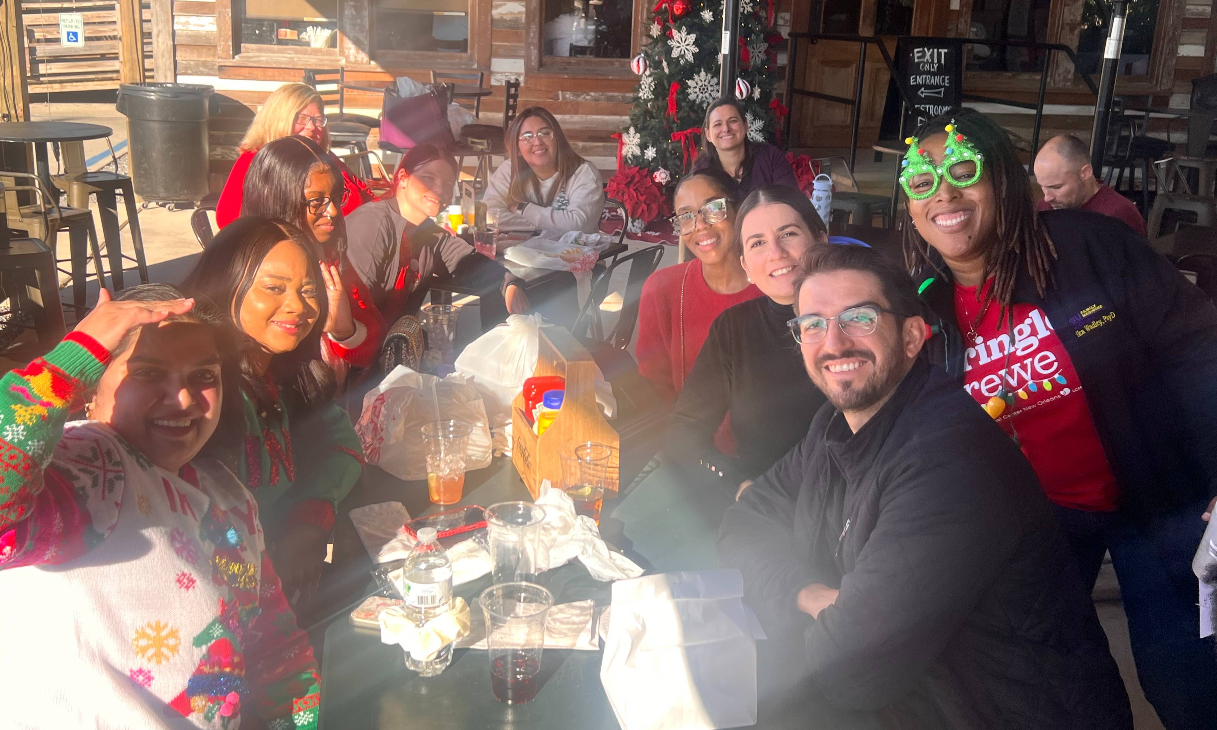 A group of colleagues gathers around tables on an outdoor restaurant patio decorated for the holiday season. Several participants wear festive sweaters, holiday-themed shirts, and novelty glasses, while food containers, drinks, and personal items are visible on the tables. A decorated Christmas tree stands near the restaurant entrance in the background, and patio heaters, chairs, and wooden building facades frame the setting. Bright sunlight creates strong highlights and lens flare across part of the image, capturing a casual holiday-themed wellness or team social event.