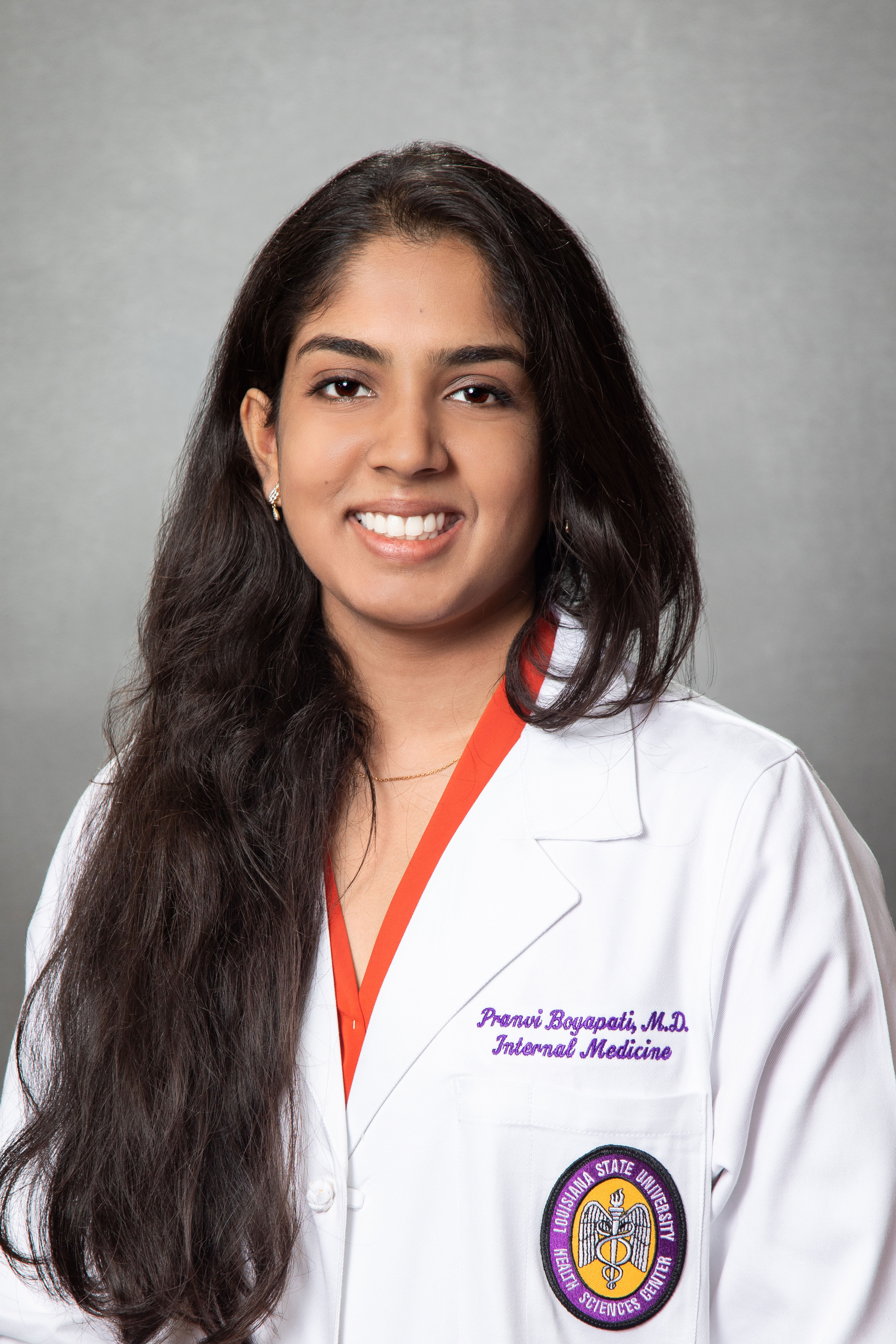 Professional medical headshot of a person wearing a white physician’s coat over a bright orange top, photographed against a gray studio background. The portrait is framed from the chest up, with long dark hair draped over one shoulder. Embroidered text on the coat reads “Pranvi Boyapati, M.D., Internal Medicine,” and a Louisiana State University Health Sciences Center patch is visible on the lower pocket. The image is lit with soft studio lighting and has a formal, clinical-profile appearance.