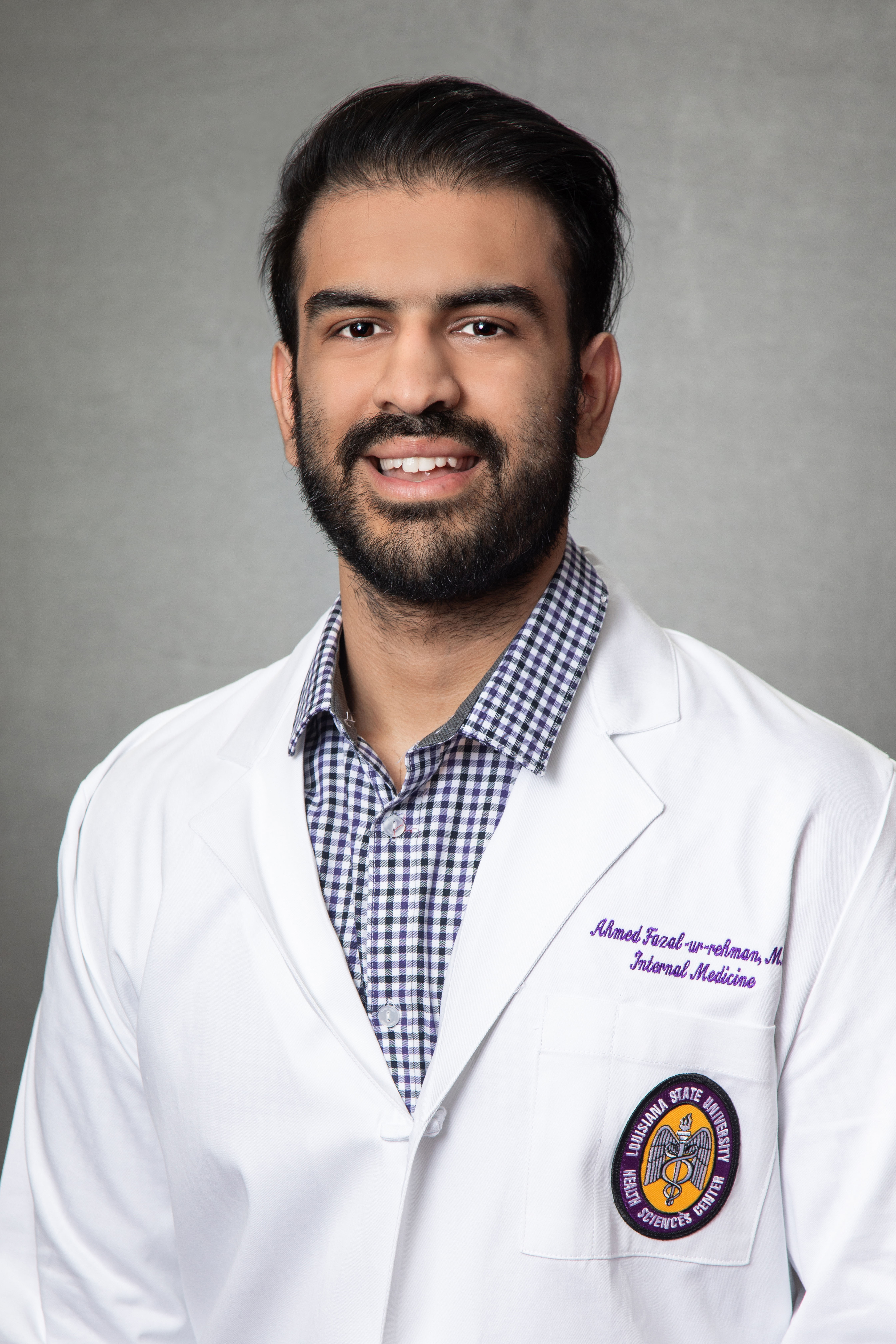 Professional medical headshot of a person wearing a white physician’s coat over a blue-and-white checkered button-down shirt, photographed against a gray studio background. The portrait is framed from the chest up, with the subject facing the camera. Embroidered text on the coat identifies the specialty as Internal Medicine, and a Louisiana State University Health Sciences Center patch is visible on the lower pocket. The image uses soft studio lighting and has a formal clinical-profile appearance.