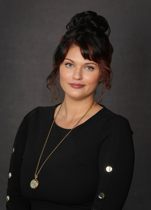 Professional headshot of an individual wearing a black top and pendant necklace against a dark studio background.
