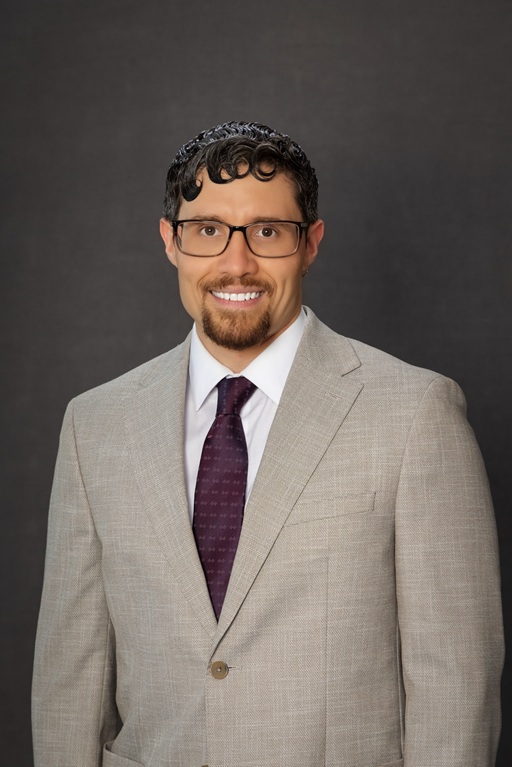 Professional headshot of an individual wearing glasses, a light gray suit, white shirt, and dark tie against a dark studio background.