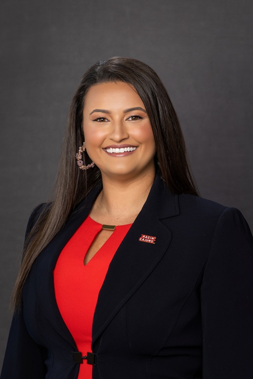 Professional headshot of a person wearing a dark blazer and red top with a Ragin’ Cajuns lapel pin against a dark studio background.