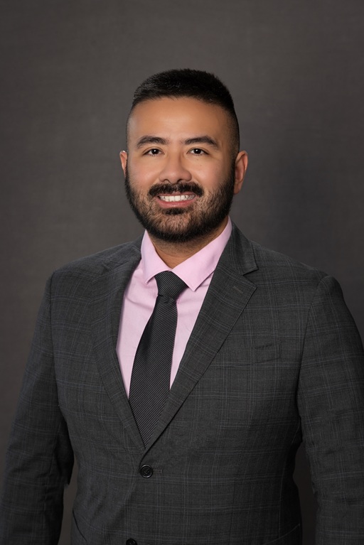 Professional headshot of an individual wearing a gray suit, pink dress shirt, and dark tie against a dark studio background.