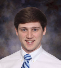 Professional headshot of a person with short brown hair, wearing a white button-down shirt and a light blue striped tie. The person is facing the camera with a closed-mouth smile. The portrait is cropped from the chest up and set against a mottled gray-and-blue studio background.