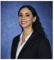 Professional headshot of a person with long dark hair pulled back, wearing a dark business jacket over a light-colored collared shirt and a small necklace. The person is posed at a slight angle toward the camera. The portrait is cropped from the chest up and set against a blue studio background.