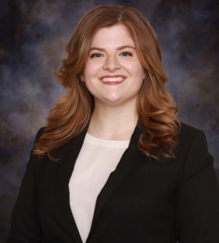 Professional headshot of a person with long, wavy, shoulder-length hair, wearing a black blazer over a light-colored blouse. The person is facing the camera. The portrait is cropped from the upper torso up and set against a mottled blue-and-gray studio background.