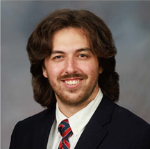 Professional headshot of a person with medium-length dark hair, wearing a dark suit jacket, white dress shirt, and red-and-blue striped tie. The person is facing the camera and smiling. The portrait is cropped from the chest up and set against a neutral gray studio background.