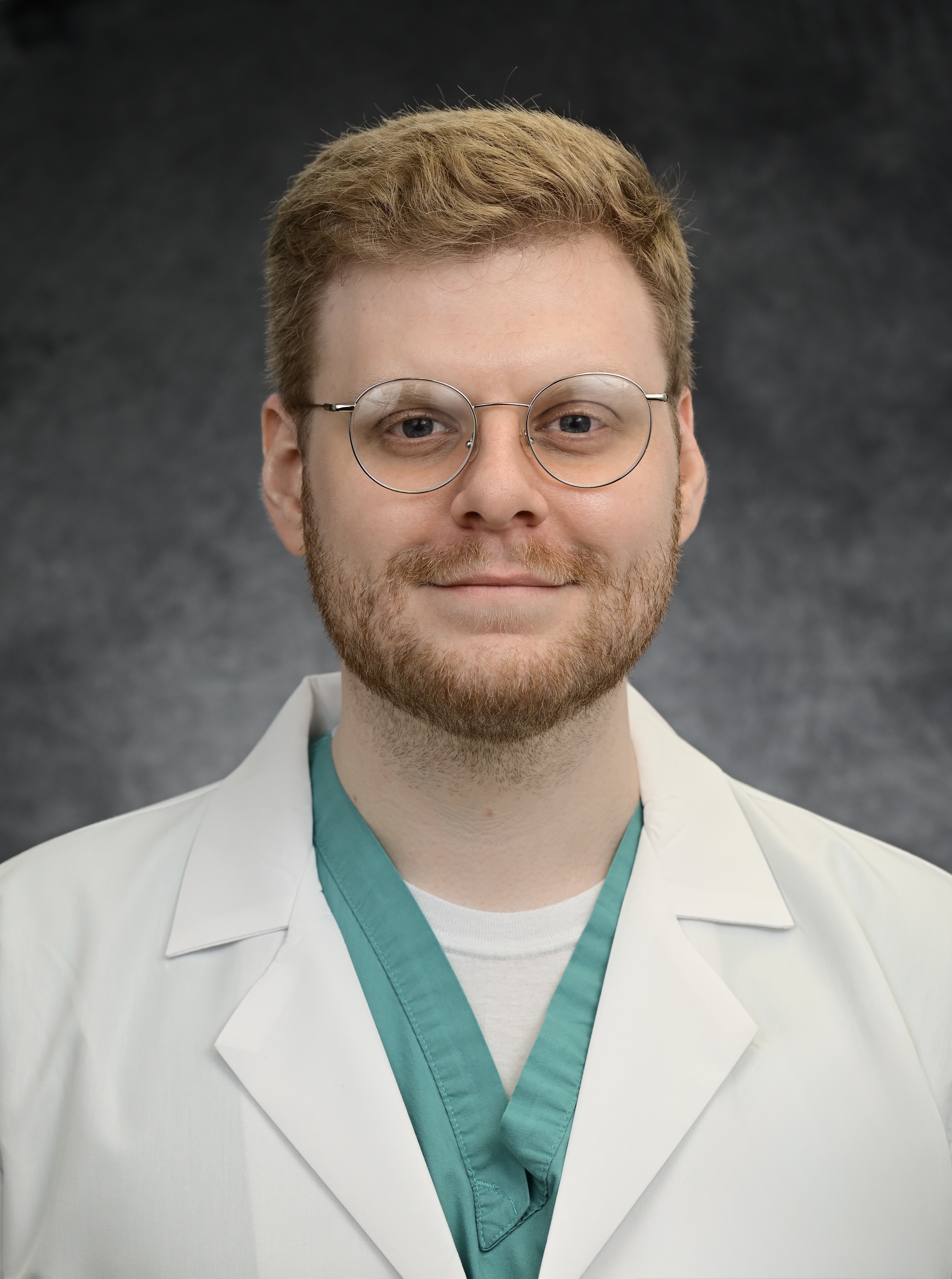Professional headshot of a person wearing round eyeglasses, a white lab coat, and teal medical scrubs. The person is facing the camera in a centered portrait against a gray studio backdrop. The image is evenly lit and cropped from the chest up.