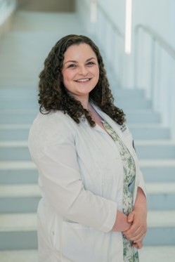 Professional portrait of an individual standing on a light-colored staircase in a bright indoor setting. The individual is wearing a light-colored coat over a patterned dress with a floral design. The portrait is framed from approximately the waist up, with hands clasped in front and the body angled slightly toward the camera. Metal handrails and softly blurred stairs create a clean, modern background with soft, even lighting and a shallow depth of field.