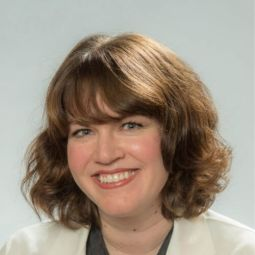 Color studio head‑and‑shoulders portrait of an adult person facing the camera against a light, neutral background. The person has short, softly curled hair and wears a light-colored blazer over a darker top. Lighting is even and soft, creating a clean, professional composition.