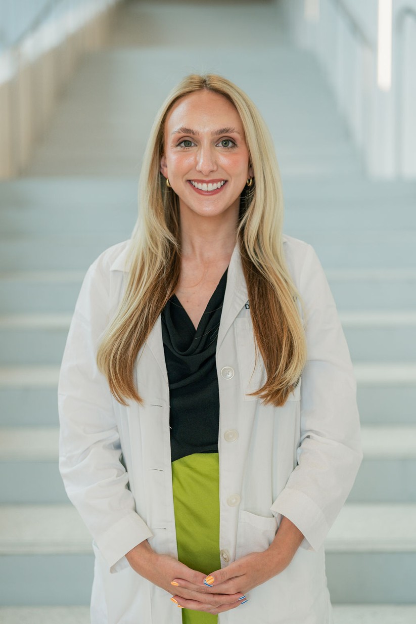 Professional portrait of an individual standing in front of a softly blurred staircase in a bright indoor setting. The individual is wearing a white medical coat over a black top and a bright green skirt. The portrait is framed from approximately the knees up, with hands clasped in front. Long light-colored hair falls past the shoulders, and small hoop earrings are visible. Light-colored stairs and metal railings create a clean, modern background with soft, even lighting and a shallow depth of field.