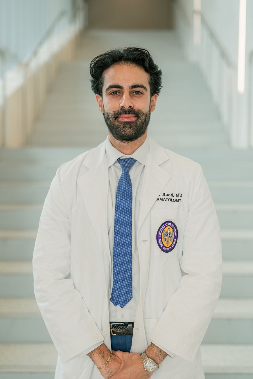 Professional portrait of an individual standing in front of a softly blurred staircase in a bright indoor setting. The individual is wearing a white medical coat over a light-colored dress shirt and a blue tie. A Louisiana State University Health Sciences Center patch is visible on the coat pocket, and embroidered text including “MD” and “Dermatology” appears on the chest. The portrait is framed from approximately the knees up, with hands clasped in front. Light-colored stairs and metal railings create a clean, modern background with soft, even lighting and a shallow depth of field.