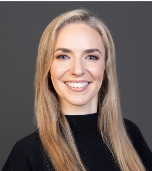 Studio portrait of a person with long, straight, light-colored hair, facing the camera against a plain gray background, wearing a black long-sleeve top; head-and-shoulders framing with even, soft lighting and teeth visible.
