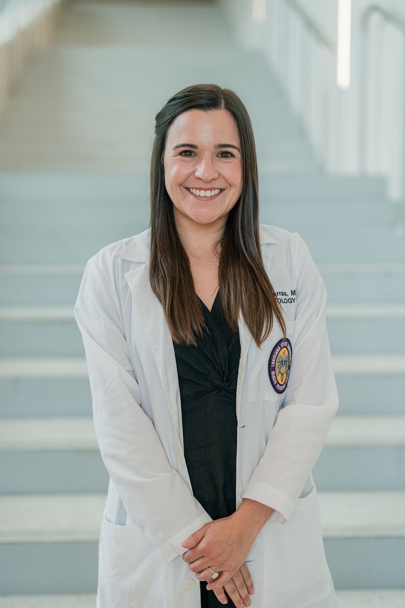 Professional portrait of an individual standing in front of a softly blurred staircase in a bright indoor setting. The individual is wearing a white medical coat over a dark V-neck outfit. A Louisiana State University Health Sciences Center patch is visible on the coat pocket, and partial embroidered text including “Dermatology” appears on the chest. The portrait is framed from approximately the waist up, with hands clasped in front. Light-colored stairs and railings create a clean, modern background with a shallow depth of field.