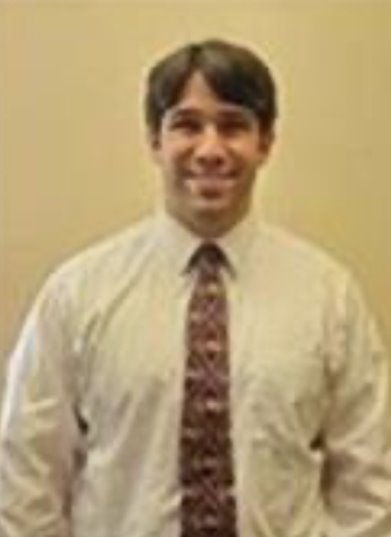 Color studio portrait of an adult person shown from the waist up, standing against a plain beige background. The person wears a light-colored button‑down shirt with a patterned tie. Hair is short and neatly styled. Lighting is soft and even, creating a simple, professional composition.