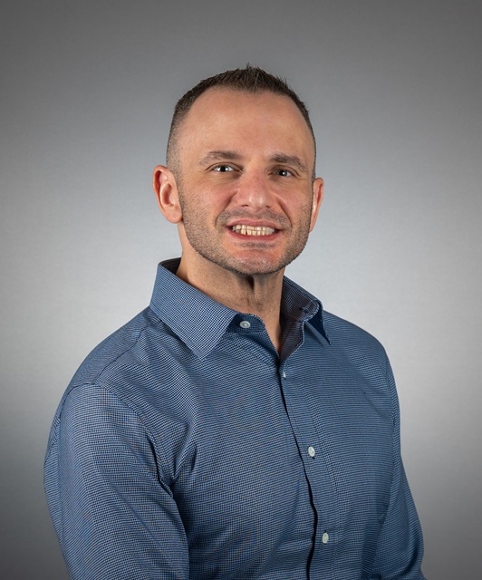 Head-and-shoulders studio portrait of a person wearing a blue long‑sleeve button‑down shirt, positioned slightly angled to the side against a plain gray background.