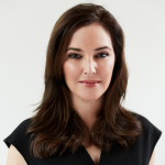 Color studio head‑and‑shoulders portrait of an adult person facing the camera against a plain white background. The person has shoulder‑length hair worn down and wears a dark, short‑sleeved top. Lighting is even and soft, creating a clean, professional composition.