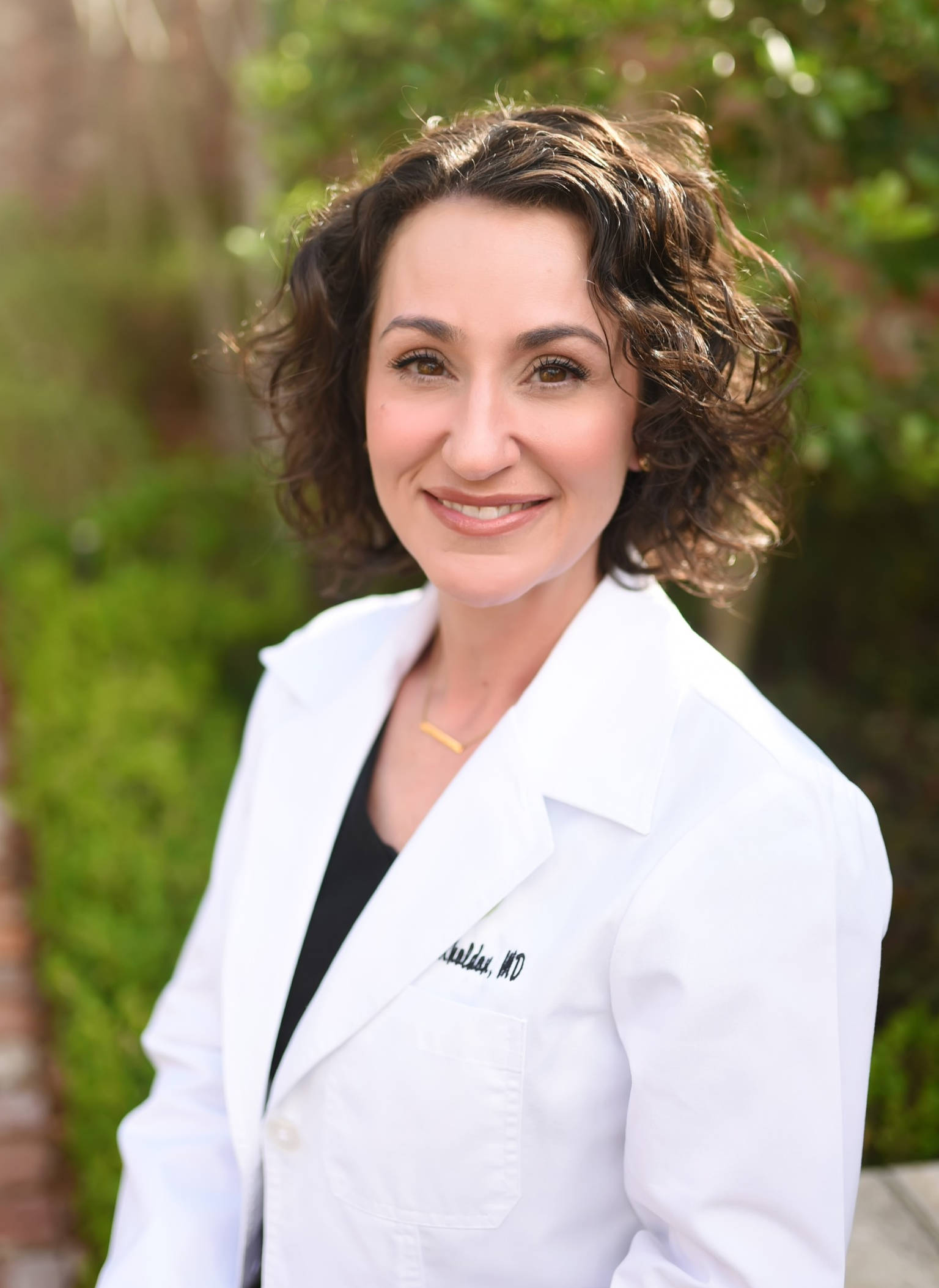 Outdoor portrait of a person with short curly hair wearing a white medical‑style coat over a dark top, seated or standing along a brick walkway bordered by greenery. Sunlight filters through plants in the softly blurred background, and embroidered text is visible on the coat.