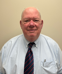 A head-and-shoulders portrait of a person standing against a plain beige background. The person is wearing a light blue button-down shirt with a dark navy tie featuring diagonal red stripes. A pen is clipped in the chest pocket of the shirt. The person has very short gray hair and is facing forward toward the camera. The lighting is even and neutral, with no visible shadows or background objects.