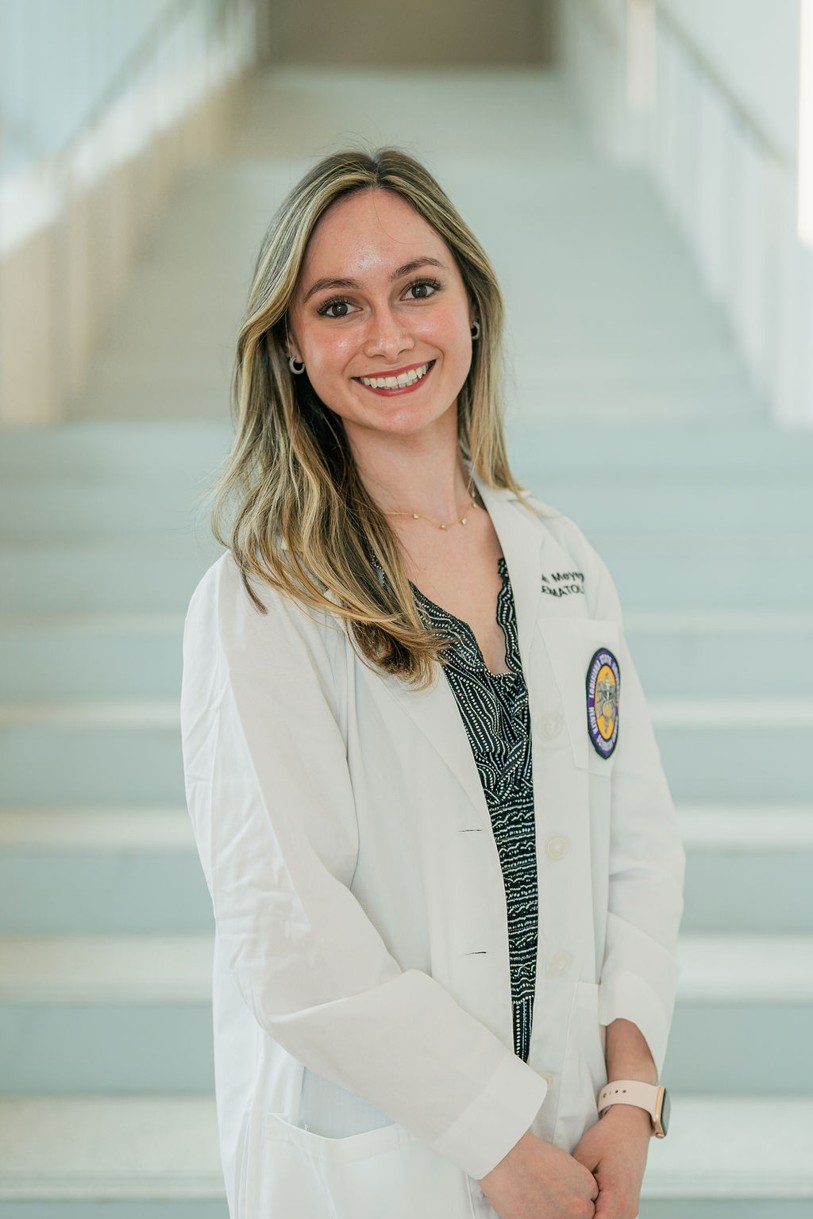 Professional portrait of an individual standing in front of a softly blurred staircase in a bright indoor setting. The individual is wearing a white medical coat over a black-and-white patterned top. A Louisiana State University Health Sciences Center patch is visible on the coat pocket, and partial embroidered text including “Dermatology” appears on the chest. The portrait is framed from approximately the knees up, with hands clasped in front. Light-colored stairs and metal railings create a clean, modern background with soft, even lighting and a shallow depth of field.