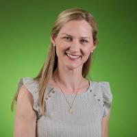 Color studio portrait of an adult person shown from the upper torso up, facing the camera against a bright green background. The person has long, straight, light-colored hair and wears a light gray sleeveless top with subtle ruffle details at the shoulders, along with a small necklace. Lighting is even and clean, creating a professional, well‑lit composition.