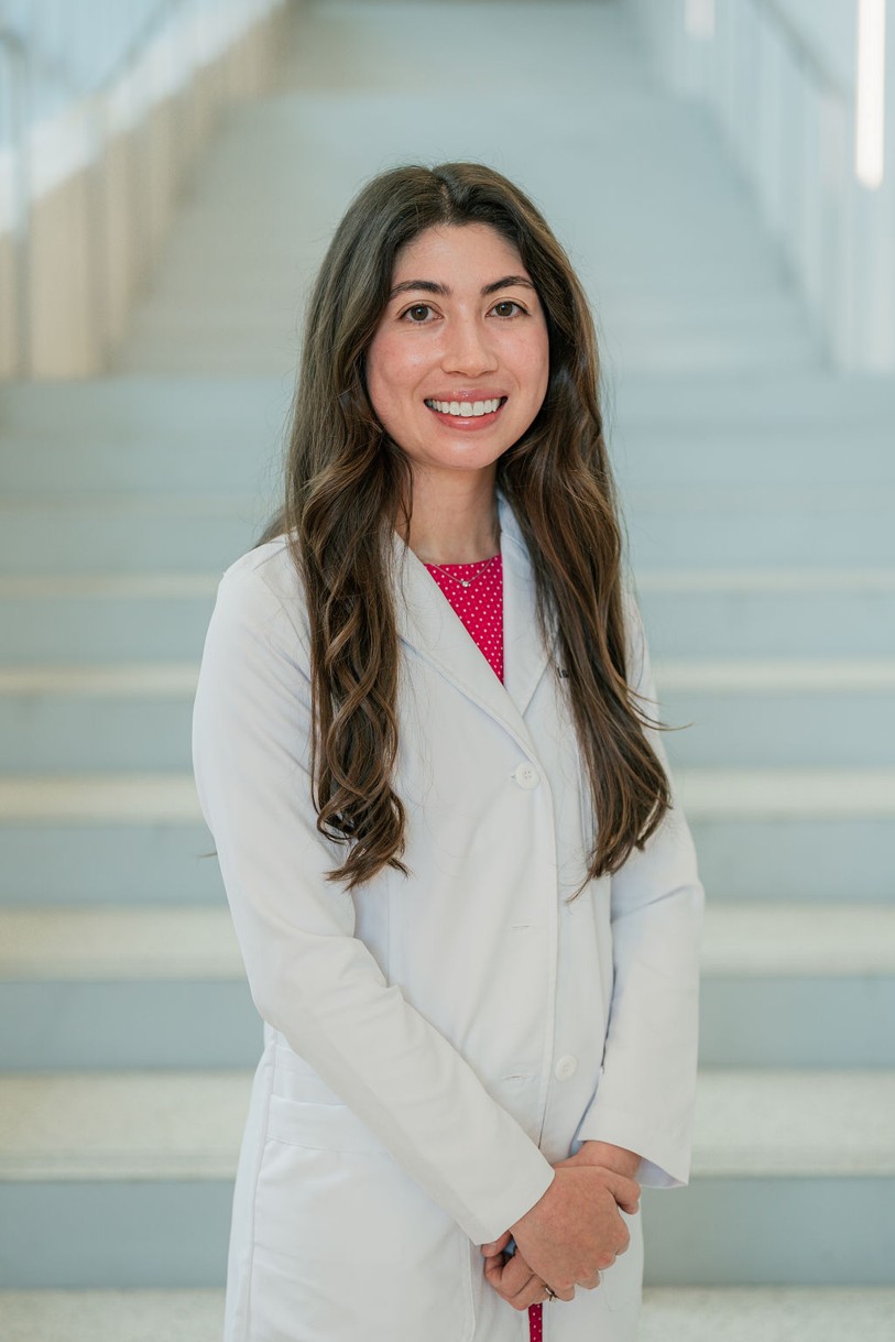Professional portrait of an individual standing in front of a softly blurred staircase in a bright indoor setting. The individual is wearing a white medical coat over a bright pink dress with a small white pattern. Long dark hair falls past the shoulders. The portrait is framed from approximately the knees up, with hands clasped in front. Light-colored stairs and metal railings create a clean, modern background with soft, even lighting and a shallow depth of field.
