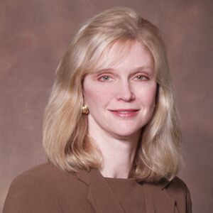 Color studio head-and-shoulders portrait of an adult person facing the camera against a softly mottled neutral background. The person has shoulder‑length light hair, wears a brown top with small earrings, and is evenly lit in a formal, professional composition.