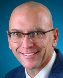 Color studio head‑and‑shoulders portrait of an adult person facing the camera against a smooth blue background. The person wears rectangular eyeglasses, a dark suit jacket, a collared shirt, and a tie. Lighting is even and professional, with a sharp, clear focus and a clean, formal composition.