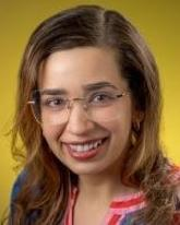 Color studio head‑and‑shoulders portrait of an adult person facing the camera against a bright yellow background. The person wears clear eyeglasses and a patterned top with red and white accents. Hair is medium length and worn down. Lighting is even and vibrant, creating a professional, friendly composition.