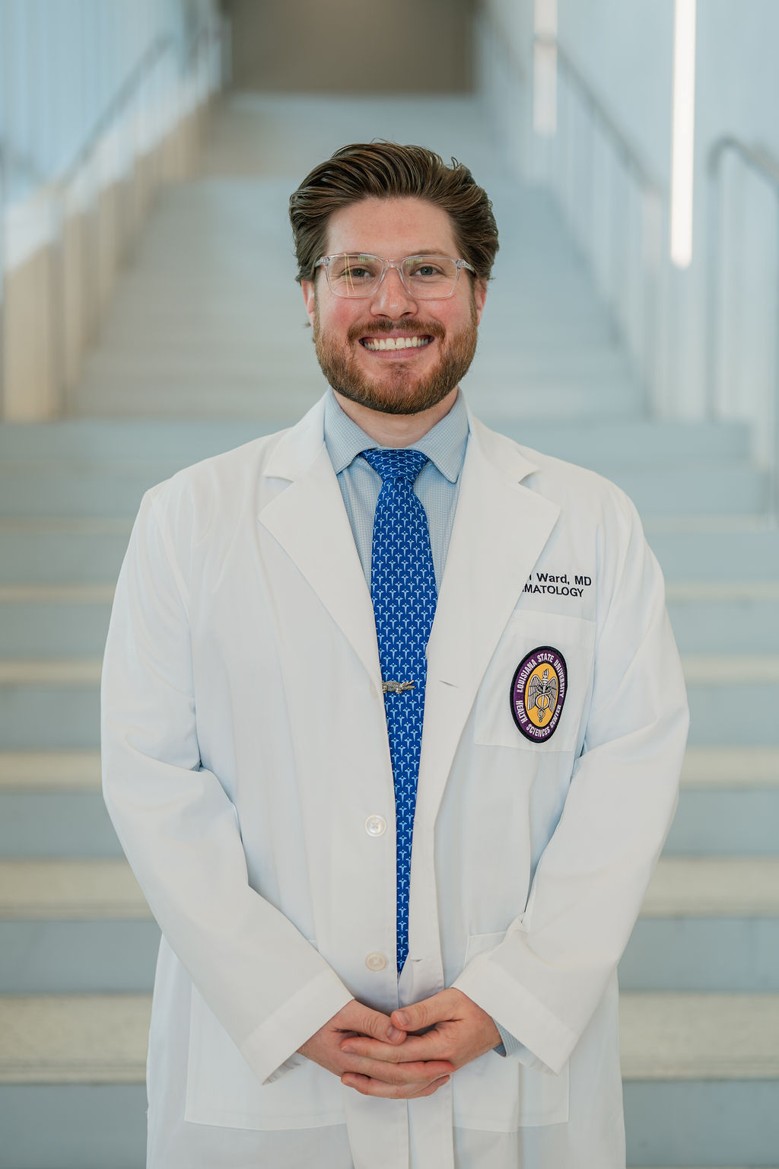 Professional portrait of an individual standing in front of a softly blurred staircase in a bright indoor setting. The individual is wearing a white medical coat over a light blue dress shirt and a blue patterned tie with a tie clip. A Louisiana State University Health Sciences Center patch is visible on the coat pocket, and partial embroidered text including “Dermatology” appears on the chest. The portrait is framed from approximately the knees up, with hands clasped in front. Light-colored stairs and railings create a clean, modern background with soft, even lighting and a shallow depth of field.
