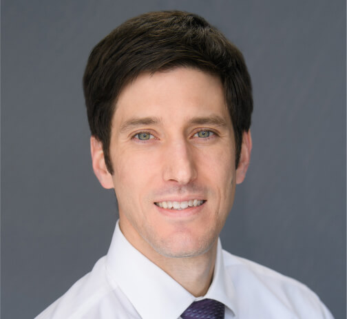 Color studio head-and-shoulders portrait of a person facing the camera against a smooth gray background. The person has short, dark hair and wears a white collared shirt with a tie. Lighting is even and soft, with a neutral, professional composition.